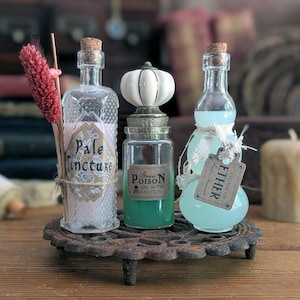 May include: Three glass bottles with cork stoppers on a round metal trivet. The left bottle contains pink liquid and is labeled Pale Tincture. The center bottle has a white pumpkin-shaped stopper and green liquid labeled Basic Poison. The right bottle contains light blue liquid and a tag labeled Essence of Ether.