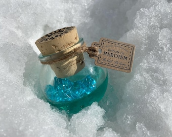 Puede incluir: Una pequeña botella de vidrio con un tapón de corcho llena de cristales azules y un líquido verde. La botella tiene una etiqueta de papel marrón adjunta que dice "Poción de Heroísmo. Sé valiente. Sé valiente."