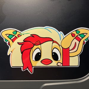 May include: A die-cut sticker featuring a cartoon character peeking out from a taco shell. The character has red hair, large eyes, and a red mouth. The taco shell is yellow with red, green, and white accents.