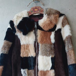 May include: A patchwork fur jacket with a brown suede body and a zip-up front. The fur is a mix of black, white, brown, and tan squares. The jacket is hanging on a wooden hanger with floral details.