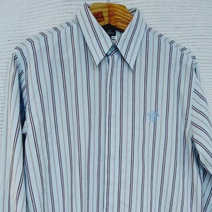 May include: A long-sleeved button-down shirt with vertical stripes in light blue, brown, and navy. The shirt has a pointed collar and a small embroidered emblem on the chest. The shirt is hanging on a wooden hanger.