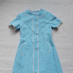 Può includere: Abito vintage azzurro con motivo floreale. L'abito presenta maniche corte, colletto smerlato e finiture bianche. Si chiude con bottoni e ha due tasche con bordi bianchi. L'abito è lungo circa 90 cm.