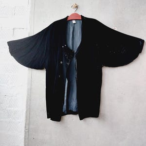 May include: A black velvet jacket with wide sleeves hangs on a wooden hanger against a grey concrete wall. The jacket has a button closure and a silky, blue-grey lining. The sleeves are flared, creating a dramatic silhouette.