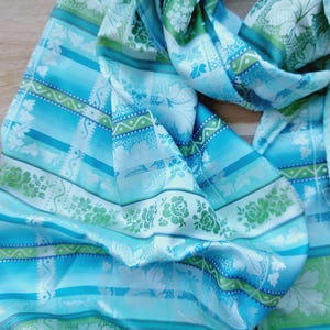 May include: A lightweight scarf featuring a repeating pattern of horizontal stripes in shades of turquoise, sky blue, and lime green. The design includes intricate white floral motifs, leaf silhouettes, and geometric borders printed on the fabric.