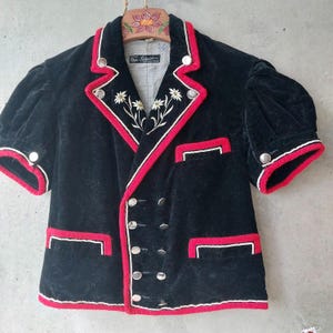May include: A black velvet short-sleeve jacket featuring red and white trim, silver-toned buttons, and white floral embroidery on the lapels. The jacket has a double-breasted front, two lower pockets, one chest pocket, and puff sleeves.