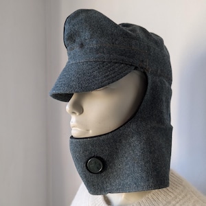 May include: A gray winter hat with a visor and face covering. The hat has a button detail on the face covering. The hat is made of a soft, felt-like material. The hat is designed to protect the face and head from the cold.