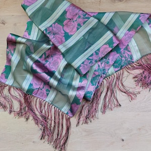 May include: A rectangular scarf featuring a pattern of pink roses on a dark green background with alternating vertical stripes in a lighter green shade. The edges of the scarf are finished with long, thin, dark pink and black fringe tassels.