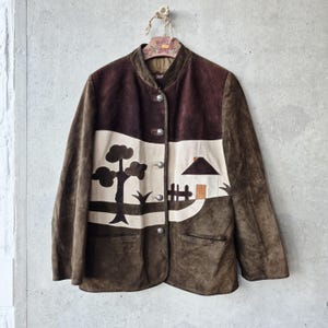 May include: A brown suede jacket featuring a cream-coloured horizontal panel across the chest. The panel displays a dark brown appliqué landscape design of a tree, a house, and a fence. The jacket has a mandarin collar and four silver-toned buttons.