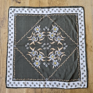 May include: A square scarf featuring a dark olive green center with a symmetrical pattern of light blue and yellow edelweiss flowers. A thin, braided rope-like border frames the center, surrounded by a white border with small floral motifs.