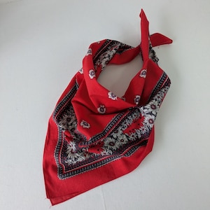 May include: A bright red bandana featuring a dark blue and white floral border pattern. The fabric is tied into a triangular shape, showcasing a central red field surrounded by a detailed decorative frame of repeating flower motifs and geometric lines.