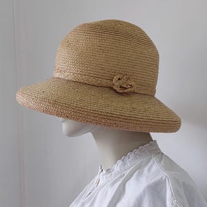 May include: A light tan straw hat with a rounded crown and a wide, slightly downturned brim. The hat features a braided straw band around the base of the crown, accented with a small, circular, woven straw flower detail on the side.