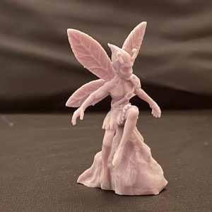 May include: A purple 3D printed figurine of a fairy with wings made of leaves. The fairy is sitting on a rock and has a detailed face and body.