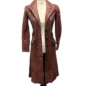 May include: A long, dark brown leather coat featuring a notched collar, long sleeves, and a front button closure. The coat includes two flap pockets on the front and decorative stitching details along the chest area, extending to the waistline.