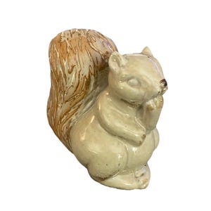 May include: A ceramic squirrel figurine in a seated pose, with a light beige body and a textured, tan-colored tail. The squirrel is holding a nut, with a brown nose and a glossy finish.