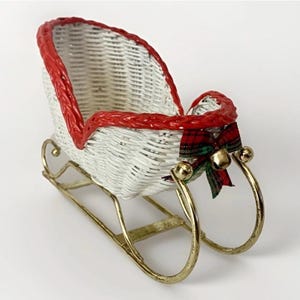 May include: A decorative sleigh featuring a white woven wicker basket body with a bright red braided rim. The sleigh is mounted on gold-toned metal runners and is accented at the front with a small plaid fabric bow and a single gold bell.