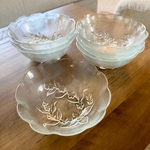 May include: A set of clear glass bowls with a scalloped edge and floral design. The bowls are stacked in groups of five and six, with one bowl in the foreground. The bowls are on a wooden surface.