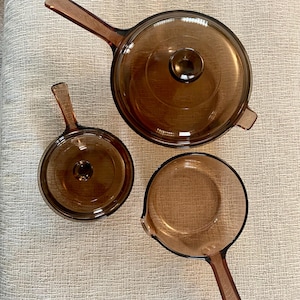 May include: A set of three brown glass cookware pieces, including a saucepan with a lid, a smaller saucepan with a lid, and a round pan with a handle and a pouring spout. The cookware has a vintage aesthetic.