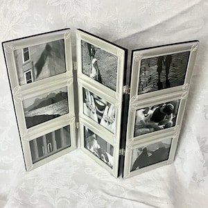 May include: A folding three-panel picture frame with a white finish and decorative leaf-like corner accents. The frame holds six rectangular black and white photographs arranged in two vertical columns of three, connected by metal hinges.