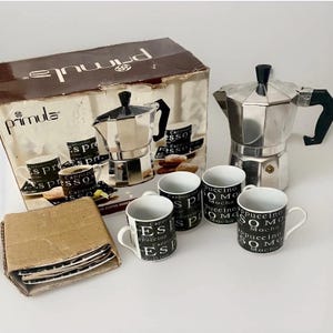 May include: A Primula brand stovetop espresso maker with a metallic silver finish and black handle, accompanied by four white ceramic mugs featuring black text patterns and a cardboard box containing matching saucers with similar text designs.
