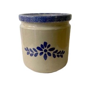 Op de afbeelding: Een beige keramische pot met een blauw bloemmotief. De pot heeft een cilindrische vorm met een blauw gespikkelde rand. Het bloemmotief is gecentreerd op de voorkant van de pot.