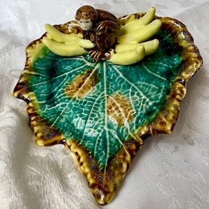 Könnte beinhalten: Eine keramikförmige Schale in Blattform mit einer Affenfigur und Bananen. Die Schale hat ein grünes und braunes Blattdesign mit einem detaillierten Adernmuster. Der Affe ist braun mit weißem Gesicht, und die Bananen sind gelb.