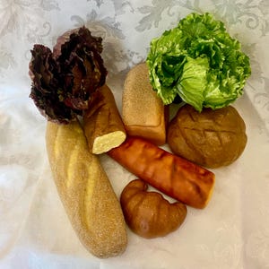 May include: A collection of artificial food items including a long baguette, a rectangular loaf, a round loaf, a croissant, a cylindrical roll, a head of bright green lettuce, and a cluster of dark purple radicchio leaves arranged together.
