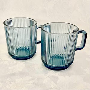 May include: Two matching mugs made of translucent blue glass, each featuring a vertical ribbed texture along the exterior walls, a cylindrical body shape, and a sturdy, integrated handle extending from the side of the vessel.