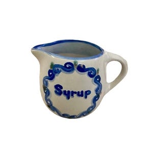 Puede incluir: Una pequeña jarra de cerámica blanca para jarabe con borde y asa azules. La palabra "Syrup" está escrita en azul dentro de un borde decorativo azul en la parte delantera de la jarra. La jarra tiene un pico.