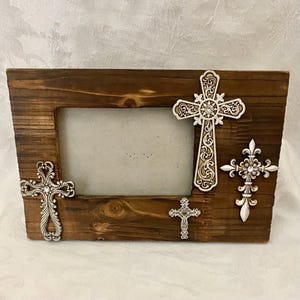 May include: A rectangular dark brown wooden picture frame featuring three silver-toned metal cross embellishments. The crosses have intricate scrollwork patterns and small embedded clear crystals. The frame has a horizontal orientation.