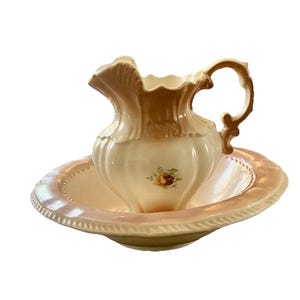 May include: A vintage porcelain pitcher and basin set. The pitcher is cream-colored with a brown rim and handle, and a floral design. The basin is a matching cream color with a decorative rim. A classic antique bathroom accessory.