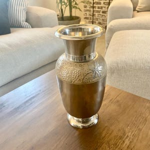 May include: A silver and bronze vase with a wide, flared rim and a round base. The upper portion of the vase is silver, while the lower portion is bronze. The bronze section features an embossed grape and leaf design.
