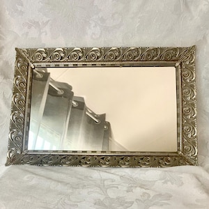 May include: A rectangular wall mirror featuring a metallic silver frame with an intricate, raised scrollwork pattern along all four edges. The inner border of the frame displays a subtle checkered detail, surrounding a clear, reflective glass surface.