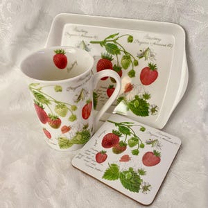 May include: A white ceramic mug, tray, and coaster set featuring a strawberry design. The set includes a mug with a handle, a rectangular tray, and a square coaster, all adorned with red and green strawberries and botanical illustrations.
