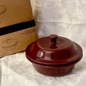 Op de afbeelding: Een diepe, ronde ovenschaal van donker kastanjebruin keramiek met een bijpassend deksel voorzien van een centrale knopgreep. De schaal staat naast een bruine kartonnen doos met de tekst One Pampered Chef Lane, Addison, Illinois 60101.
