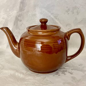 May include: A glossy, medium-brown ceramic teapot featuring a rounded body, a curved spout, a sturdy handle, and a matching lid with a small knob handle. The surface has a smooth, reflective finish with subtle horizontal ridges around the center.