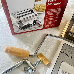 May include: A chrome-plated pasta machine with a wooden handle, a clamp, and a crank. The box reads "My Perfect Kitchen Pasta Machine" and includes text about making fresh pasta. The machine measures 7.875 inches wide.