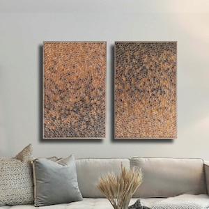 May include: Two abstract wall art pieces with a textured, speckled design in shades of brown and black. The artwork hangs above a light-coloured sofa with decorative cushions. A modern living room setting with a glass coffee table and a floor lamp.