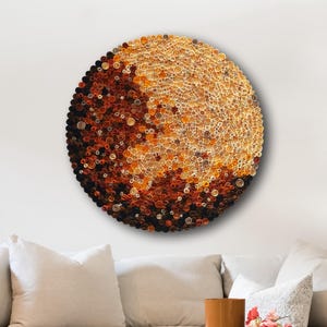 Op de afbeelding: Een groot, cirkelvormig kunstwerk aan de muur, met een kleurengradient van donkerbruin naar lichtoranje. Het stuk is gemaakt van vele kleine, getextureerde componenten. Het hangt boven een beige bank in een woonkamer.