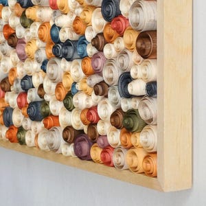 May include: A framed artwork featuring a dense arrangement of rolled wood shavings. The shavings are in various colors, including white, orange, blue, green, and brown. The wooden frame is a light, natural color.