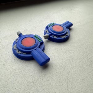 May include: Two circular blue plastic devices featuring a central red disc, a protruding handle, and small grey gear-like accents. Each device includes a row of four small green rectangular buttons and a grey cylindrical knob on the side.