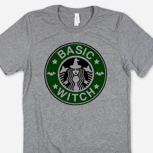 May include: A heather gray t-shirt featuring a green and black graphic. The graphic is a parody of a popular coffee shop logo, with the words "BASIC WITCH" encircling a witch illustration. The shirt is made of a soft material.