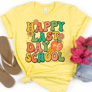 May include: Yellow t-shirt with the text "Happy Last Day of School" in colorful retro-style letters. The shirt is folded and laid flat on a white surface with a pair of brown flip-flops, a pair of blue denim shorts, and a pair of sunglasses.