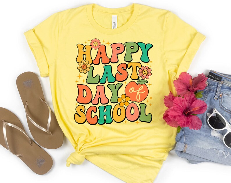 May include: Yellow t-shirt with the text "Happy Last Day of School" in colorful retro-style letters. The shirt is folded and laid flat on a white surface with a pair of brown flip-flops, a pair of blue denim shorts, and a pair of sunglasses.