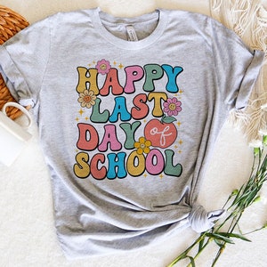 May include: A gray t-shirt with the text "Happy Last Day of School" in colorful retro-style lettering. The text is surrounded by small flower and star graphics.