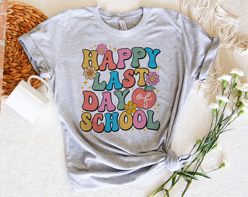 May include: A gray t-shirt with the text "Happy Last Day of School" in colorful retro-style lettering. The text is surrounded by small flower and star graphics.