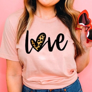 May include: A pink t-shirt with the word "love" written in black script. The "o" in love is replaced with a heart-shaped graphic with a leopard print pattern.