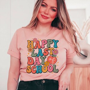 May include: A pink t-shirt with the text "Happy Last Day of School" in colorful retro letters and a red apple graphic.