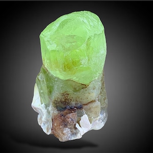 May include: A bright green, translucent, faceted crystal with a rough, brown base. The crystal is a natural specimen and has a unique, irregular shape.