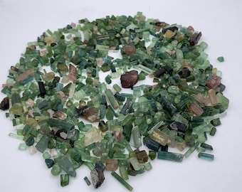 May include: A large collection of raw tourmaline crystals in various shapes and sizes. The stones display a range of colors including shades of green, light pink, brown, and black, with some pieces appearing translucent and others opaque.