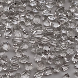Op de afbeelding: Een verzameling van heldere kwartskristallen, ook wel Herkimer-diamanten genoemd, in verschillende vormen en maten. De kristallen zijn verspreid over een wit oppervlak.
